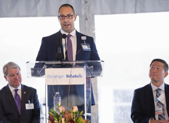 Gabriel Kamarousky speaks at a podium bearing the Geisinger and St. Luke's logos during a formal healthcare event.