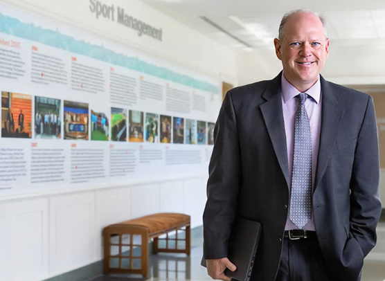 Dr. Rodney Paul stands in front of a Sport Management timeline display at Syracuse University in a professional portrait.