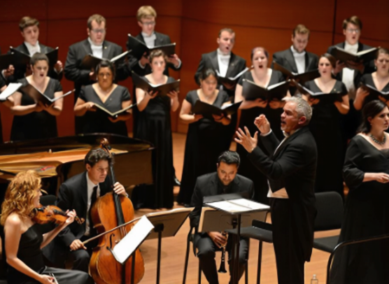 James Jordan conducts a chamber orchestra and choir during a formal performance in a wood-paneled concert hall.